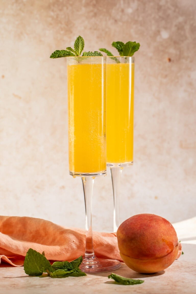 Two champagne flutes filled with peach Bellinis, garnished with fresh mint sprigs, beside a whole peach