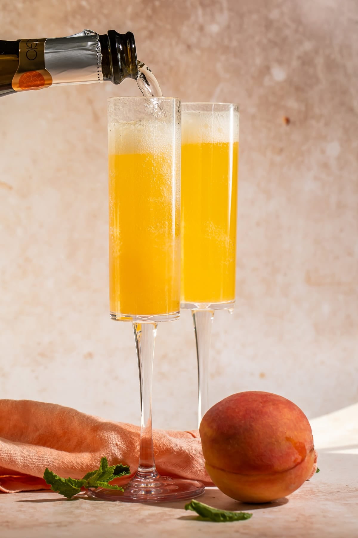 Prosecco being poured from a bottle into two champagne flutes filled with fresh peach puree