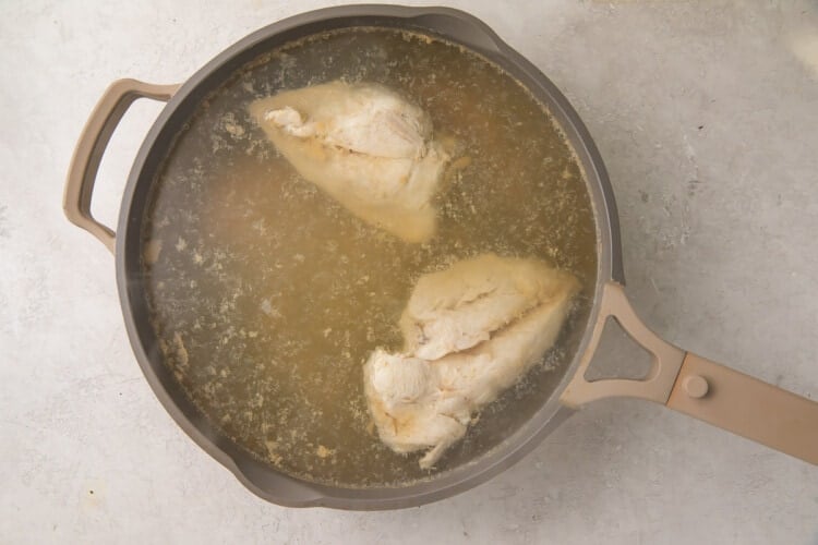 Cooked chicken breasts in chicken stock in a silver skillet