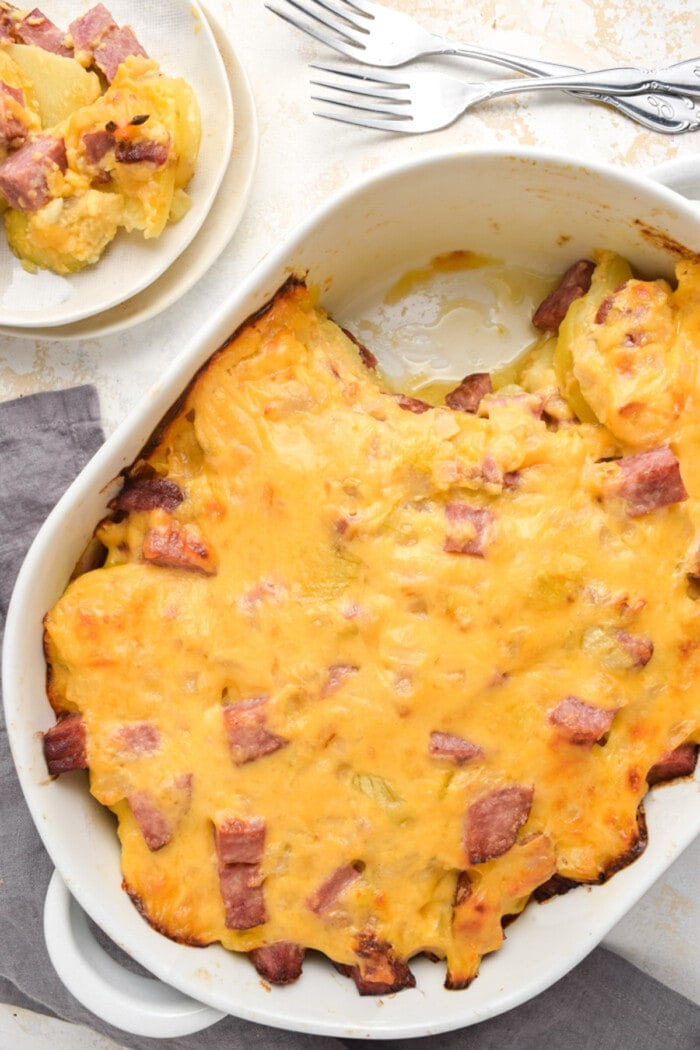 Overhead view of a casserole dish full of ham and potato casserole, with one scoop transferred to a plate.