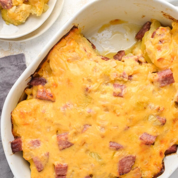 Overhead view of a casserole dish full of ham and potato casserole, with one scoop transferred to a plate.
