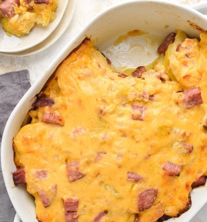 Overhead angle of cheesy ham and potato casserole in a white casserole dish, with one serving scooped onto a white plate