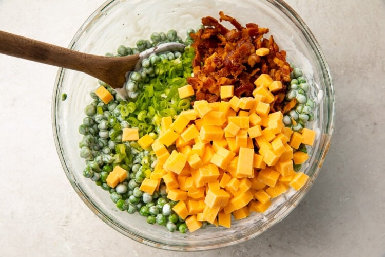 Peas, cheddar, scallions, and bacon in a large glass mixing bowl with a wooden spoon