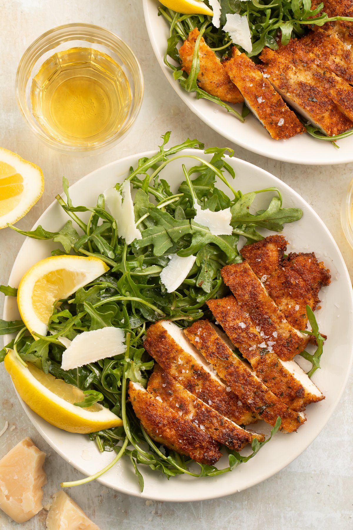 Top-down look at chicken milanese, sliced and served on a plate with a green arugula salad, lemon wedges, and parmesan slivers.
