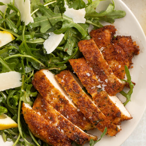 Top-down look at chicken milanese, sliced and served on a plate with a green arugula salad, lemon wedges, and parmesan slivers.