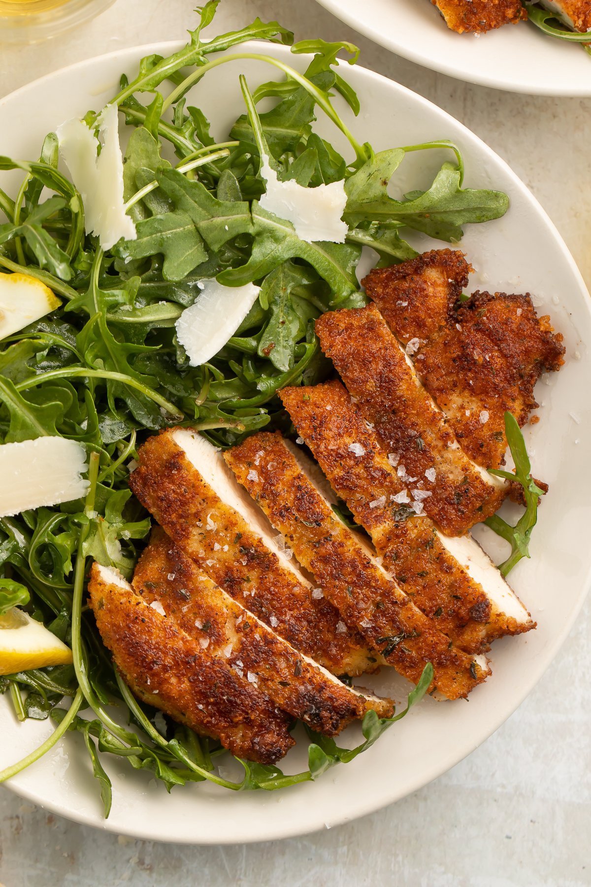 Top-down look at chicken milanese, sliced and served on a plate with a green arugula salad, lemon wedges, and parmesan slivers.