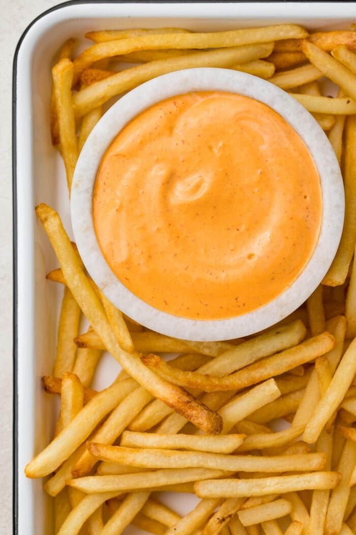 A small marble bowl of chipotle aioli surrounded by french fries on a baking sheet