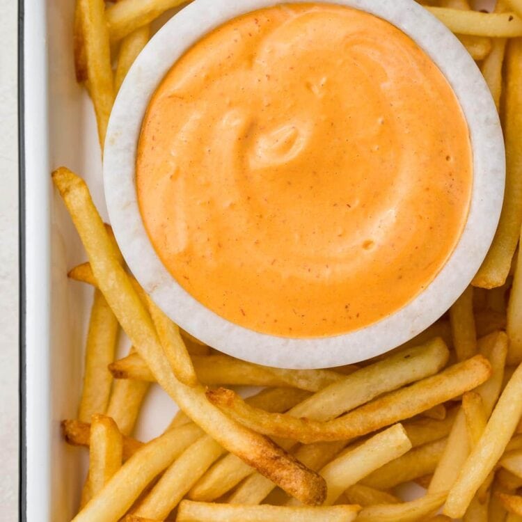 A small marble bowl of chipotle aioli surrounded by french fries on a baking sheet