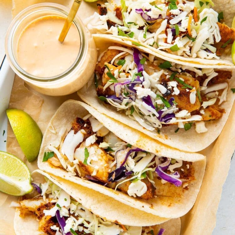 4 fish tacos lined up on a platter next to a glass jar of fish taco sauce