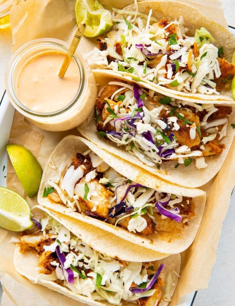 4 fish tacos lined up on a platter next to a glass jar of fish taco sauce