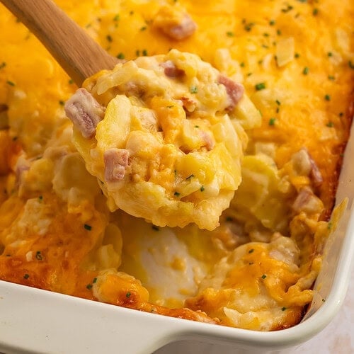A spoonful of cheesy ham and potato casserole being held over a casserole dish.