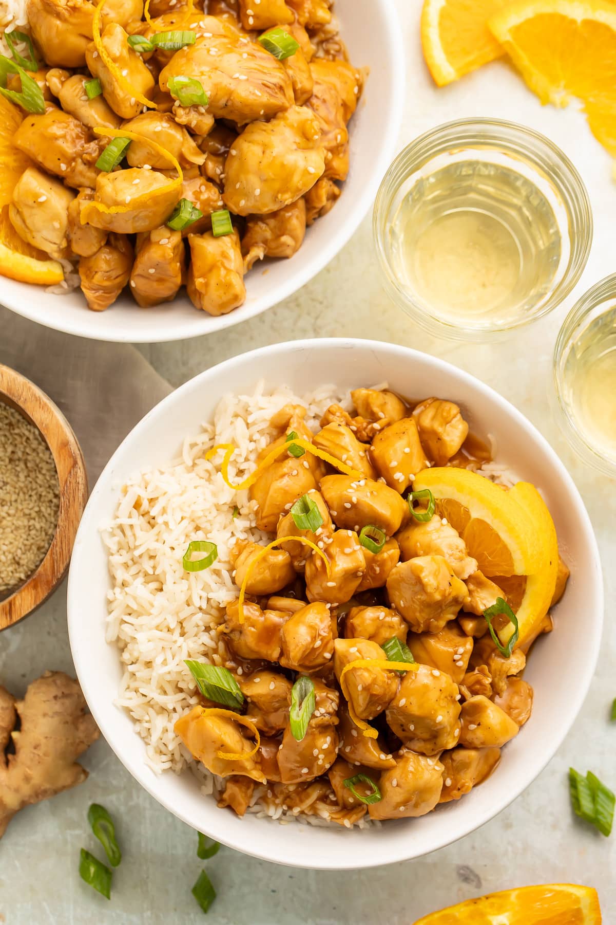 Overhead view of 2 bowls holding fluffy white rice, pieces of glazed orange chicken, orange wedges, and thinly sliced green onions.