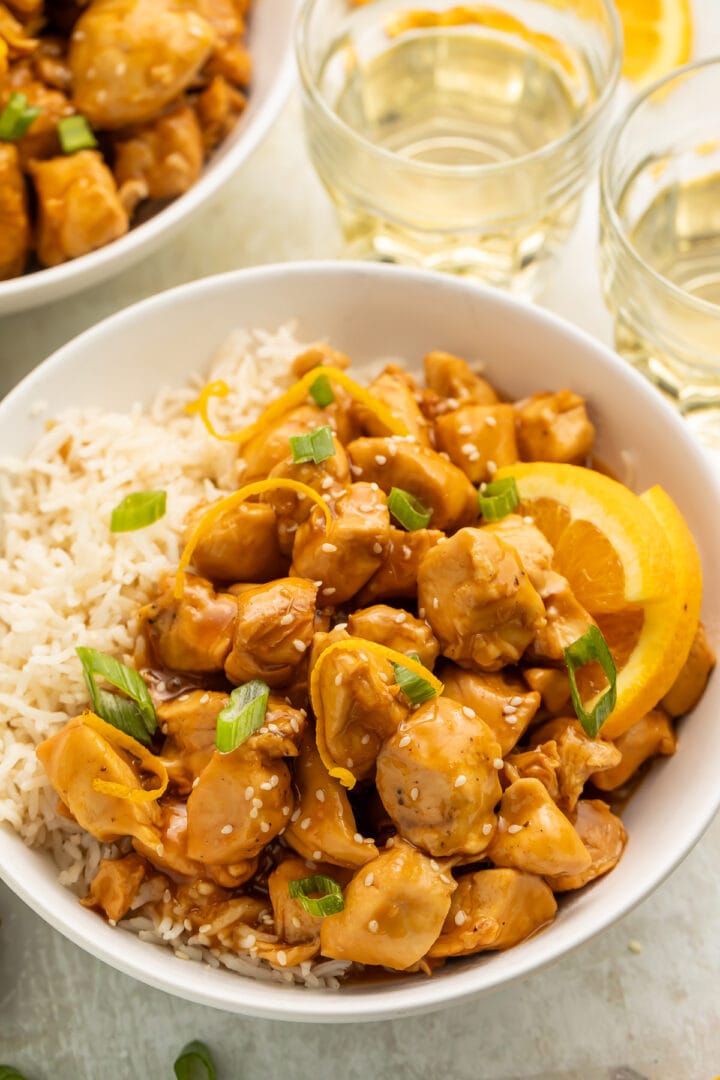 A white bowl filled with white rice, glazed orange chicken, wedges of oranges, and sliced green onions, on a table with glasses of water.