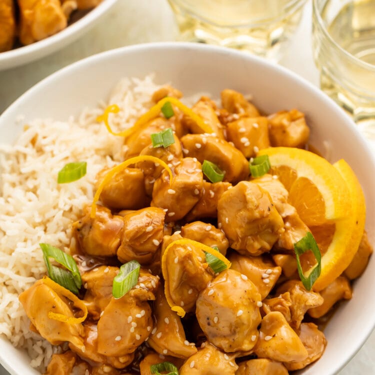 A white bowl filled with white rice, glazed orange chicken, wedges of oranges, and sliced green onions, on a table with glasses of water.