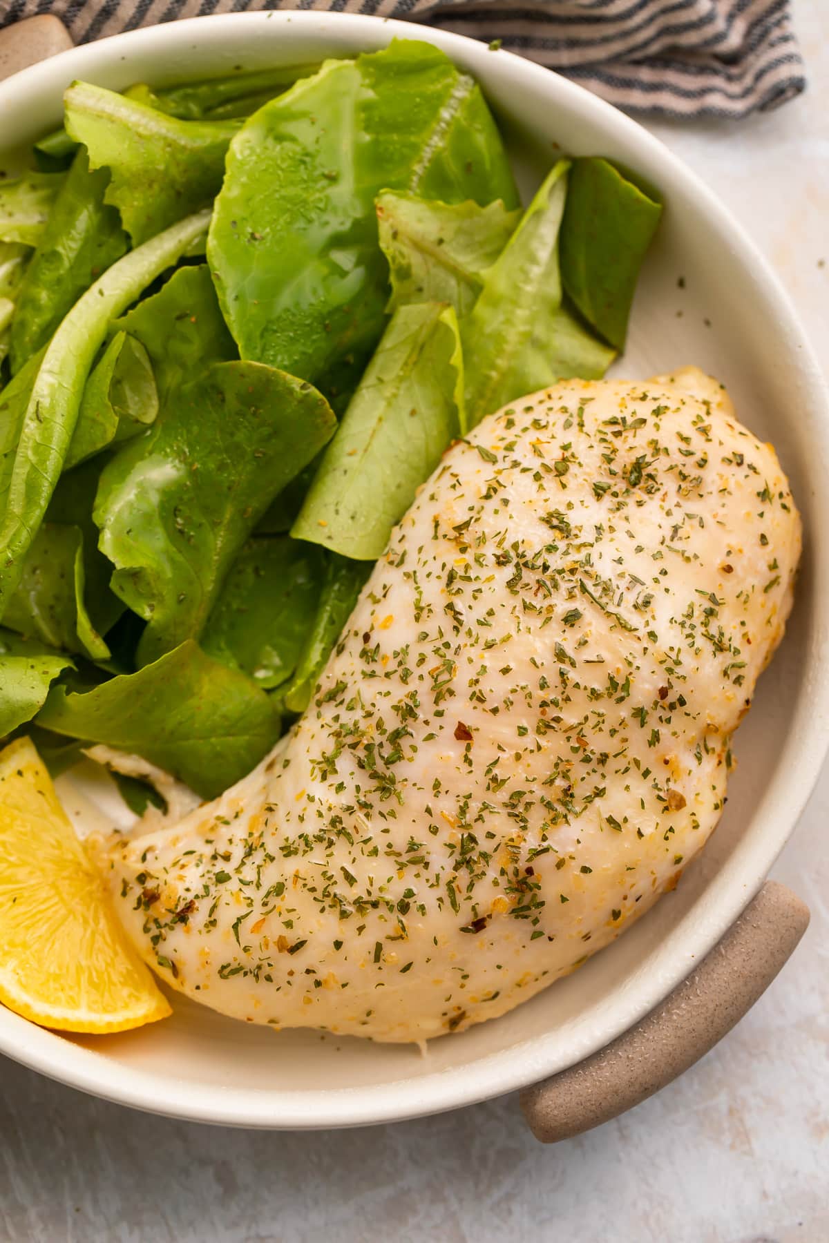 An Italian dressing chicken breast in a shallow bowl with a spinach side salad and a lemon wedge.