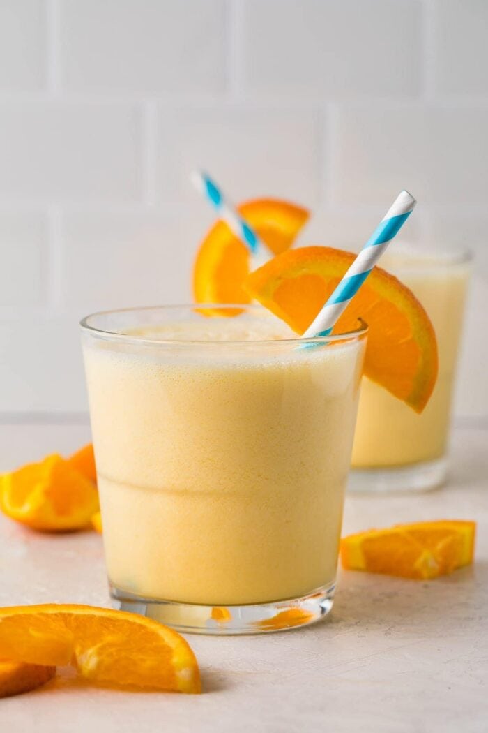 Short glass containing a serving of Orange Julius with an orange slice and a blue and white striped straw