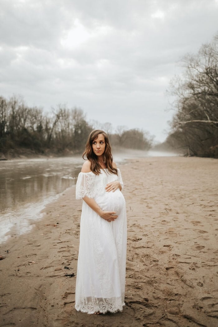 Pregnant woman in a white maxi dress on a sandy riverbank with overcast sky