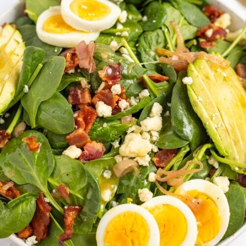 Spinach Salad with Hot Bacon Dressing in a bowl topped with hard boiled eggs, avocado, gorgonzola cheese, and crispy bacon.