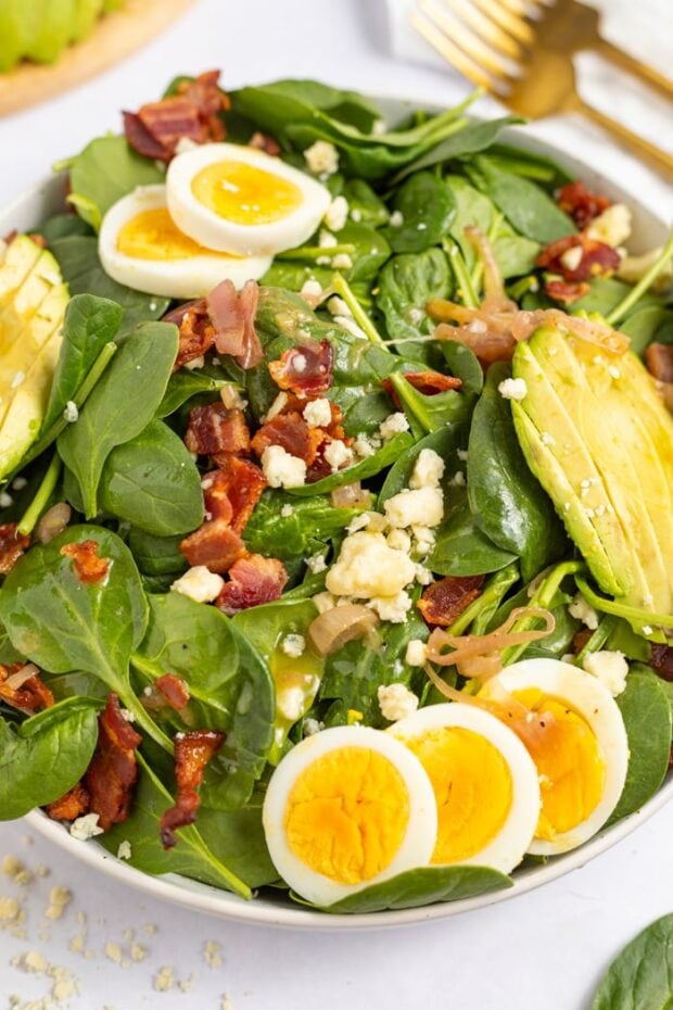 Spinach Salad with Hot Bacon Dressing in a bowl topped with hard boiled eggs, avocado, gorgonzola cheese, and crispy bacon.