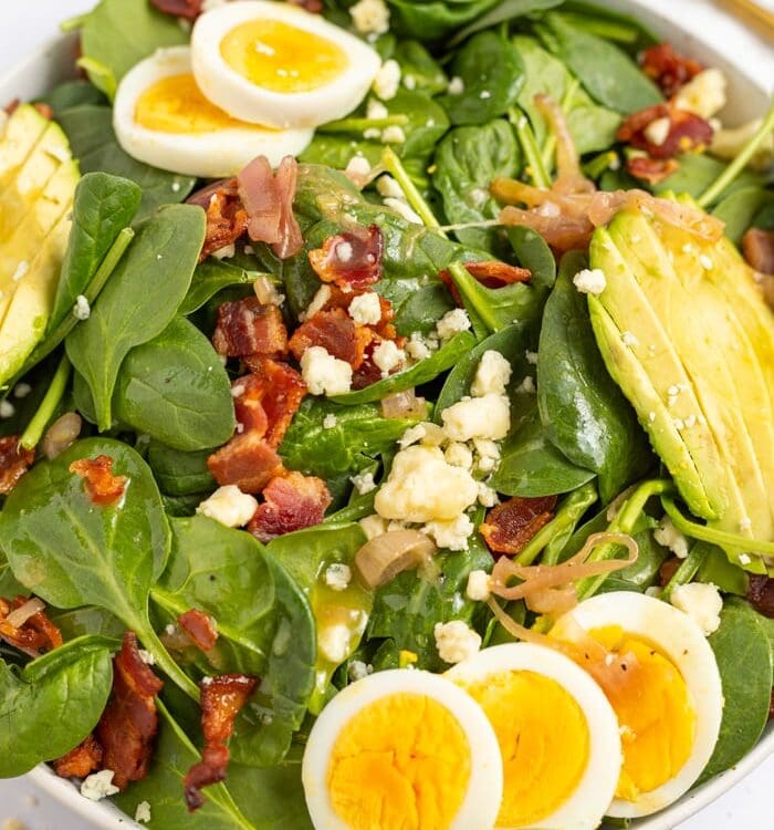 Spinach Salad with Hot Bacon Dressing in a bowl topped with hard boiled eggs, avocado, gorgonzola cheese, and crispy bacon.