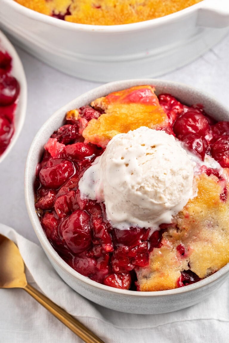 A bowl of cherry cobbler topped with a scoop of vanilla ice cream sitting on a counter next to a baking dish of cherry cobbler.