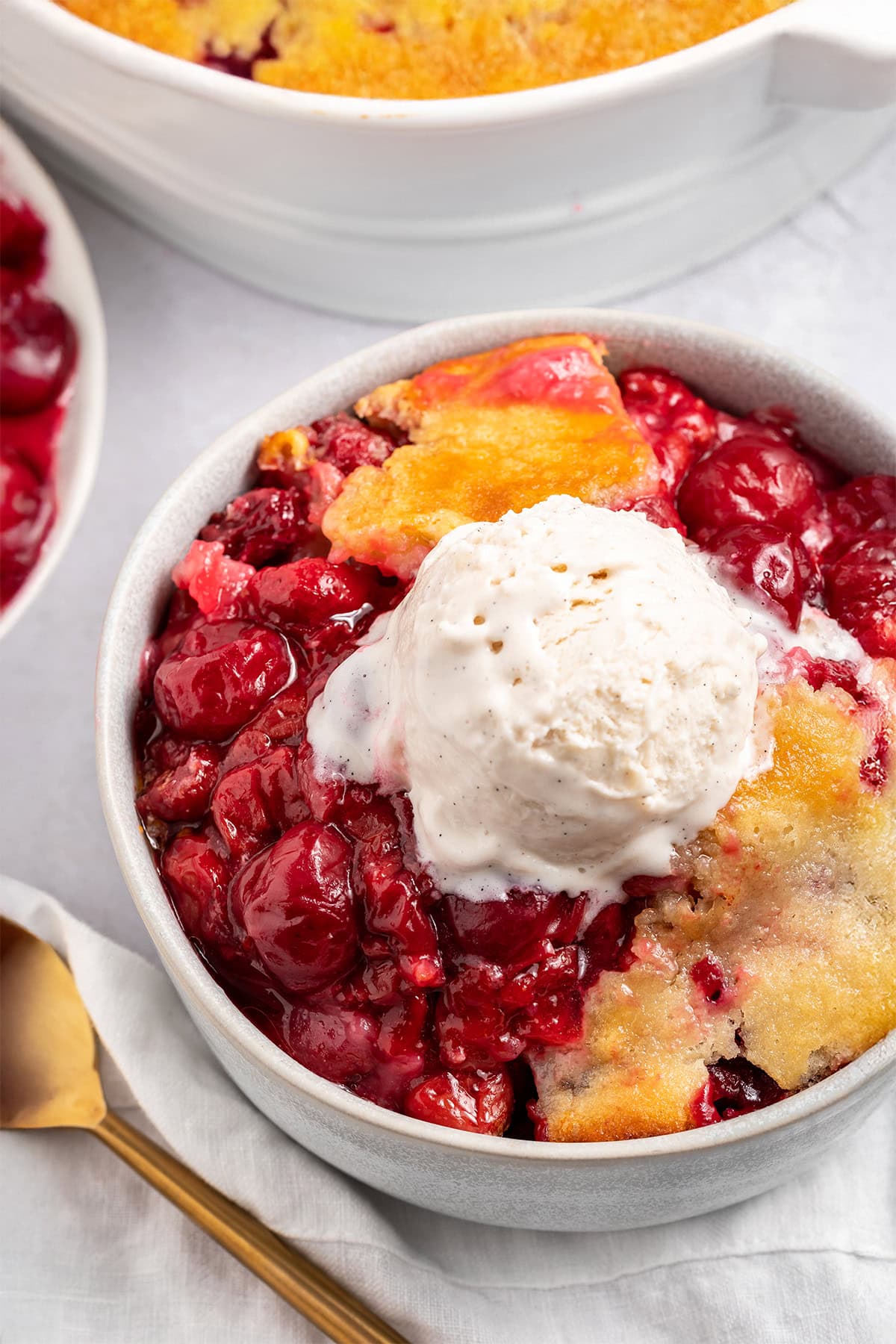 A bowl of cherry cobbler topped with a scoop of vanilla ice cream sitting on a counter next to a baking dish of cherry cobbler.