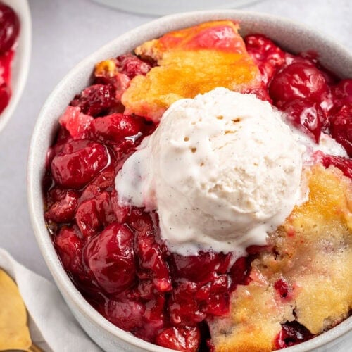 A bowl of cherry cobbler topped with a scoop of vanilla ice cream sitting on a counter next to a baking dish of cherry cobbler.