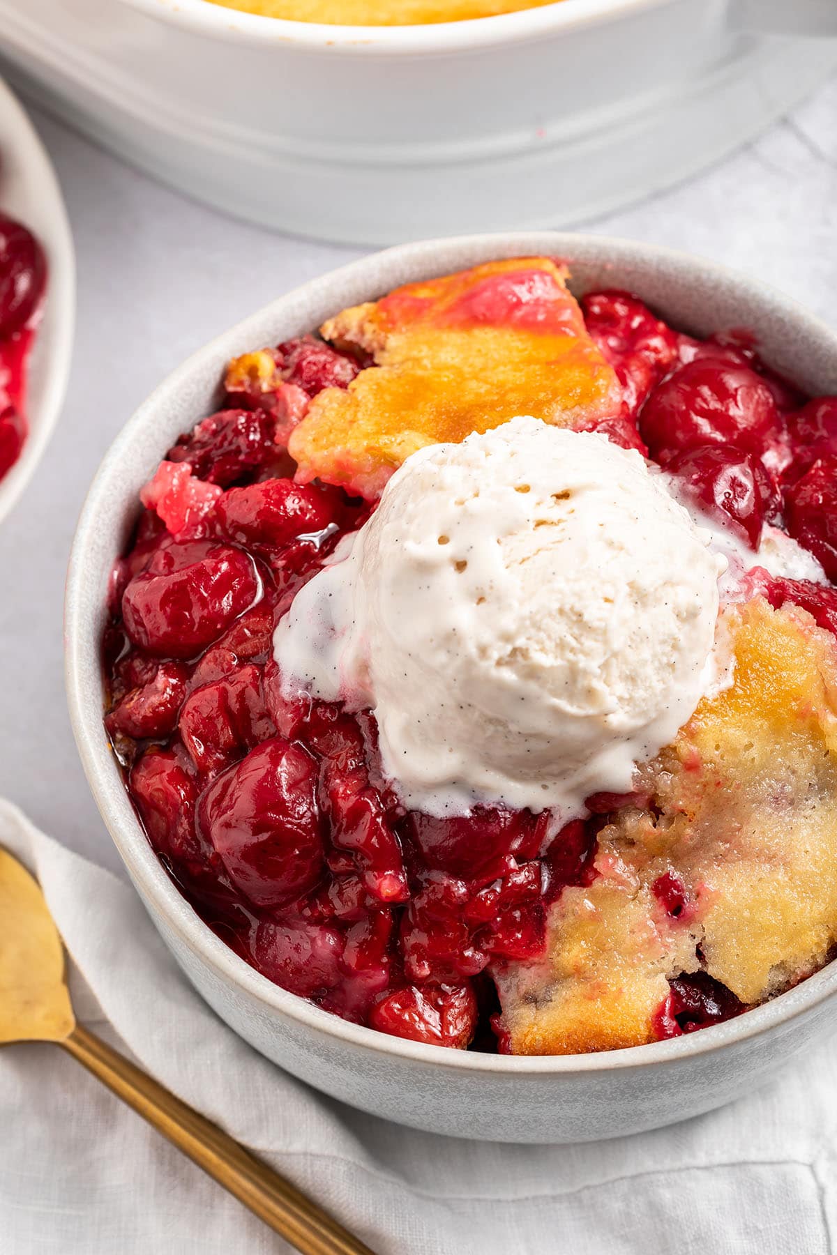 A bowl of cherry cobbler topped with a scoop of vanilla ice cream sitting on a counter next to a baking dish of cherry cobbler.