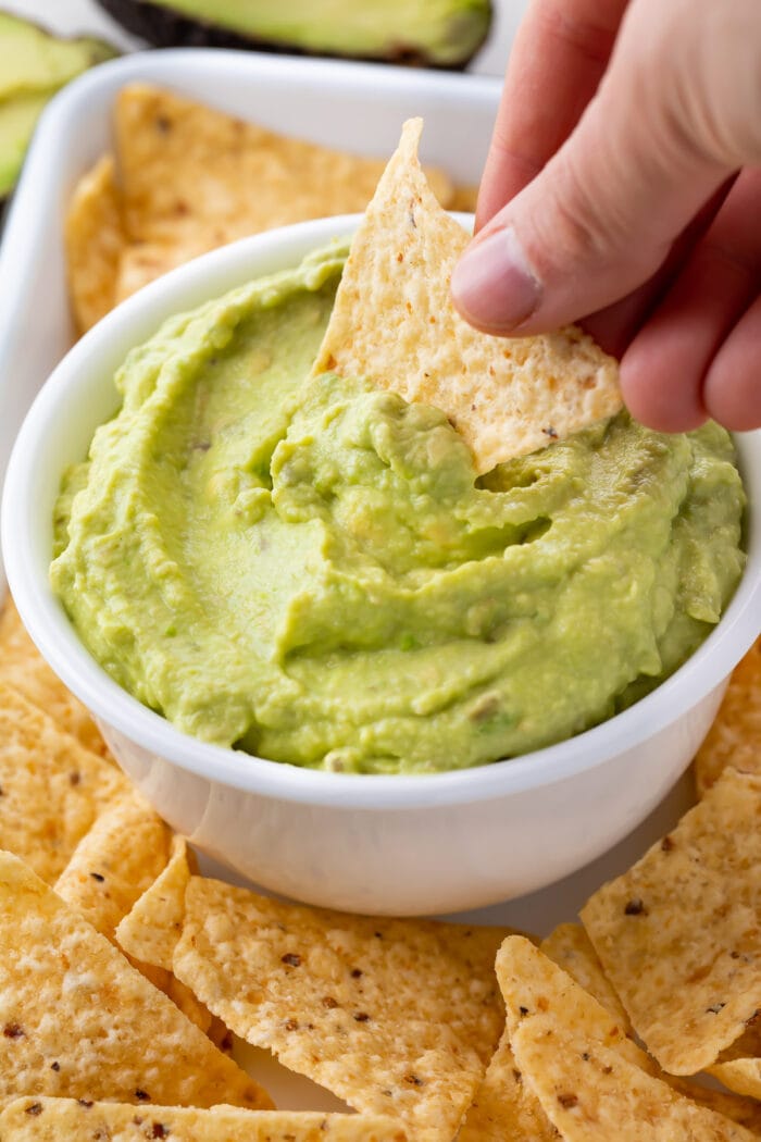 A hand dipping a tortilla chip into a bowl of guacamole dip surrounded by other tortilla chips
