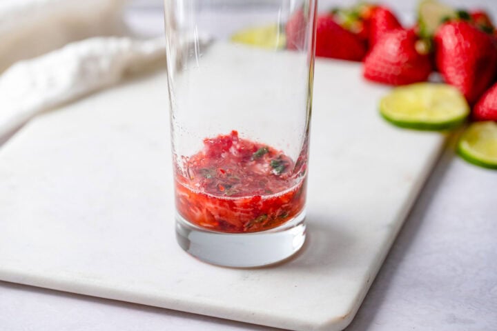 Muddled strawberries and mint leaves in the bottom of a highball glass.