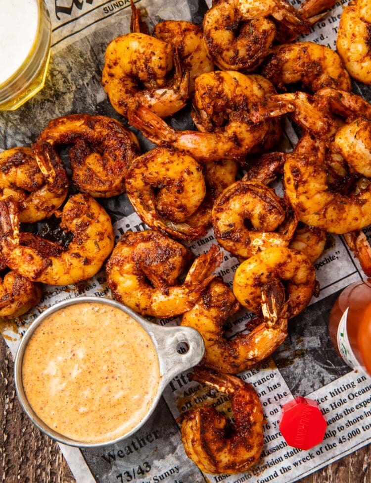 A pile of blackened shrimp on newspaper next to a dipping cup of remoulade sauce
