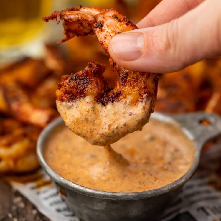 A single blackened shrimp being dipped into a small bowl of remoulade