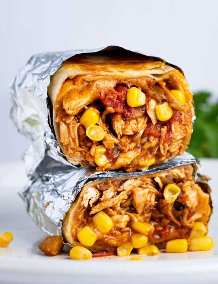 Two chicken burritos stacked on top of each other, cut crosswise to show chicken, corn, refried beans, and tomatoes inside