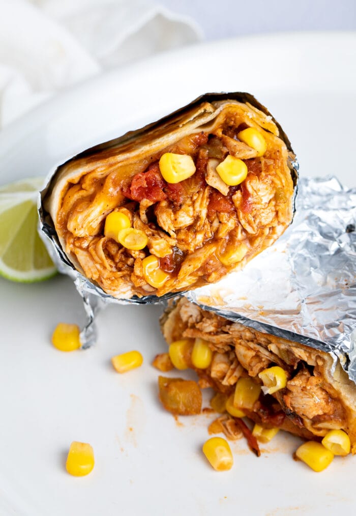 Two chicken burritos stacked on top of each other at an angle, cut crosswise to show chicken, corn, refried beans, and tomatoes inside