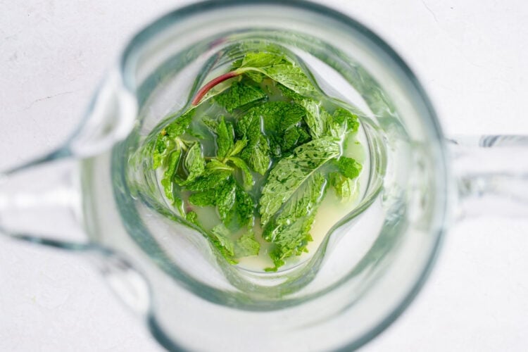 Muddled mint leaves and fresh lime juice in the bottom of a glass pitcher.