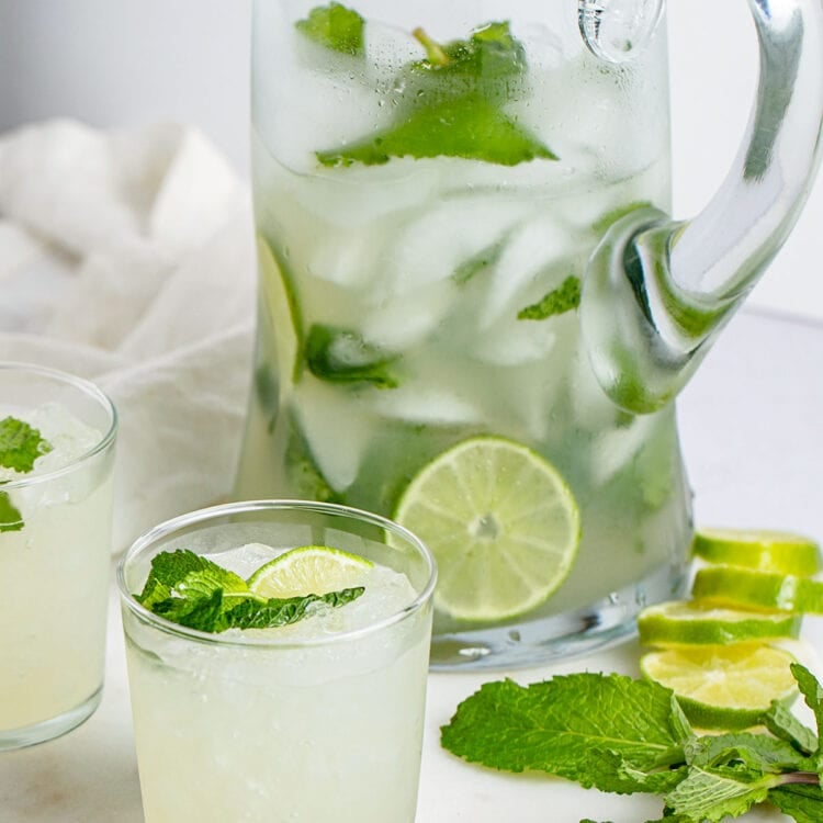 A mojito in a short glass sits in front of a tall glass pitcher of mojitos.