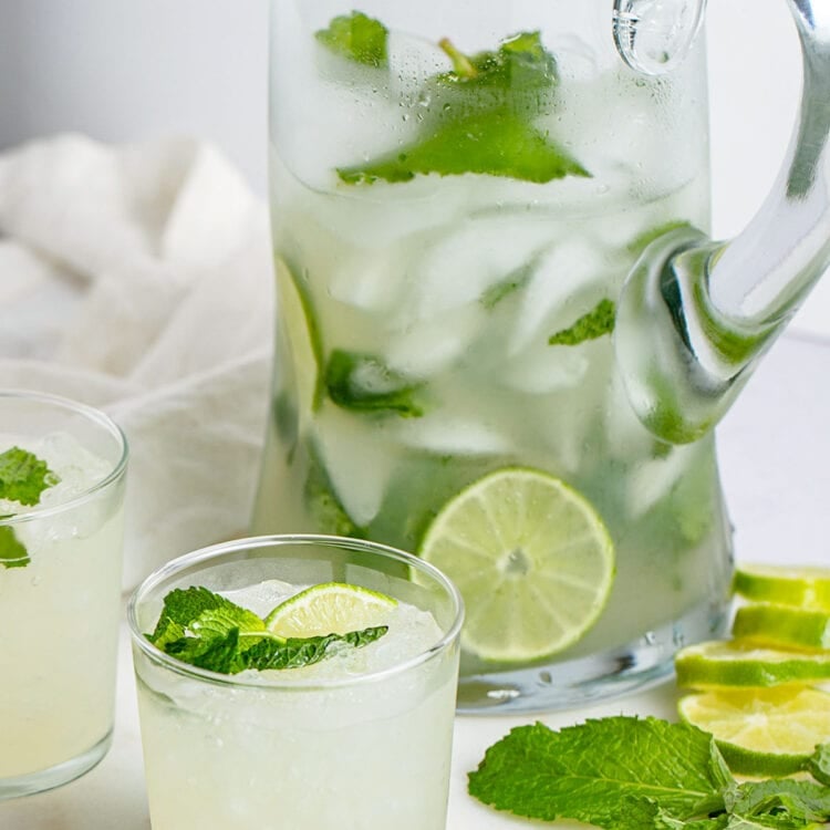A mojito in a short glass sits in front of a tall glass pitcher of mojitos.