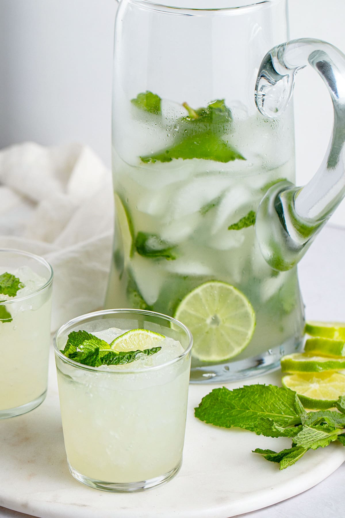 A mojito in a short glass sits in front of a tall glass pitcher of mojitos.