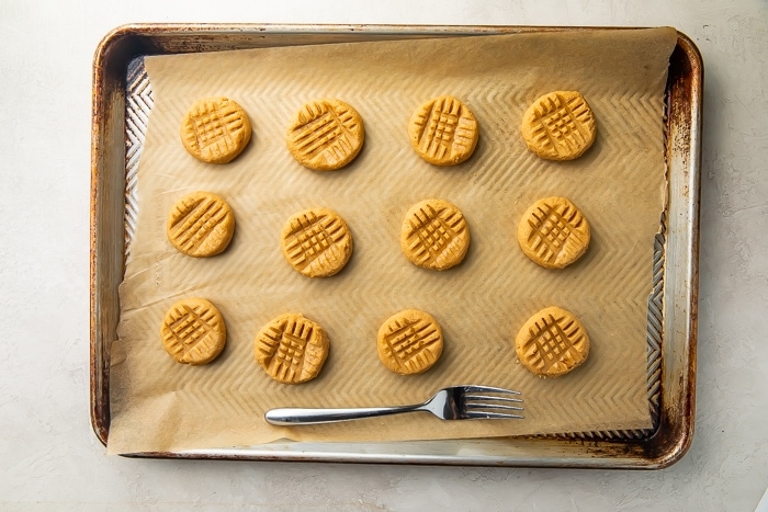 Gluten free peanut butter cookies on baking sheet lined with parchment paper next to fork used to make criss-cross pattern on top of cookies