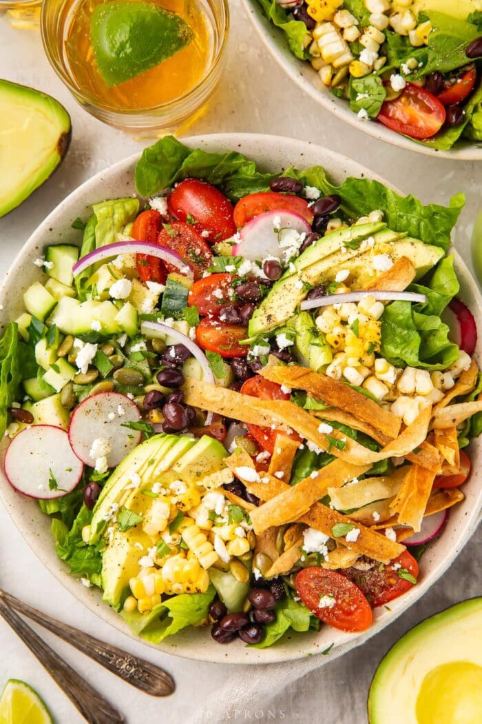 Overhead angle of Mexican salad with tortilla strips in a white bowl, next to a glass of chelada and limes.