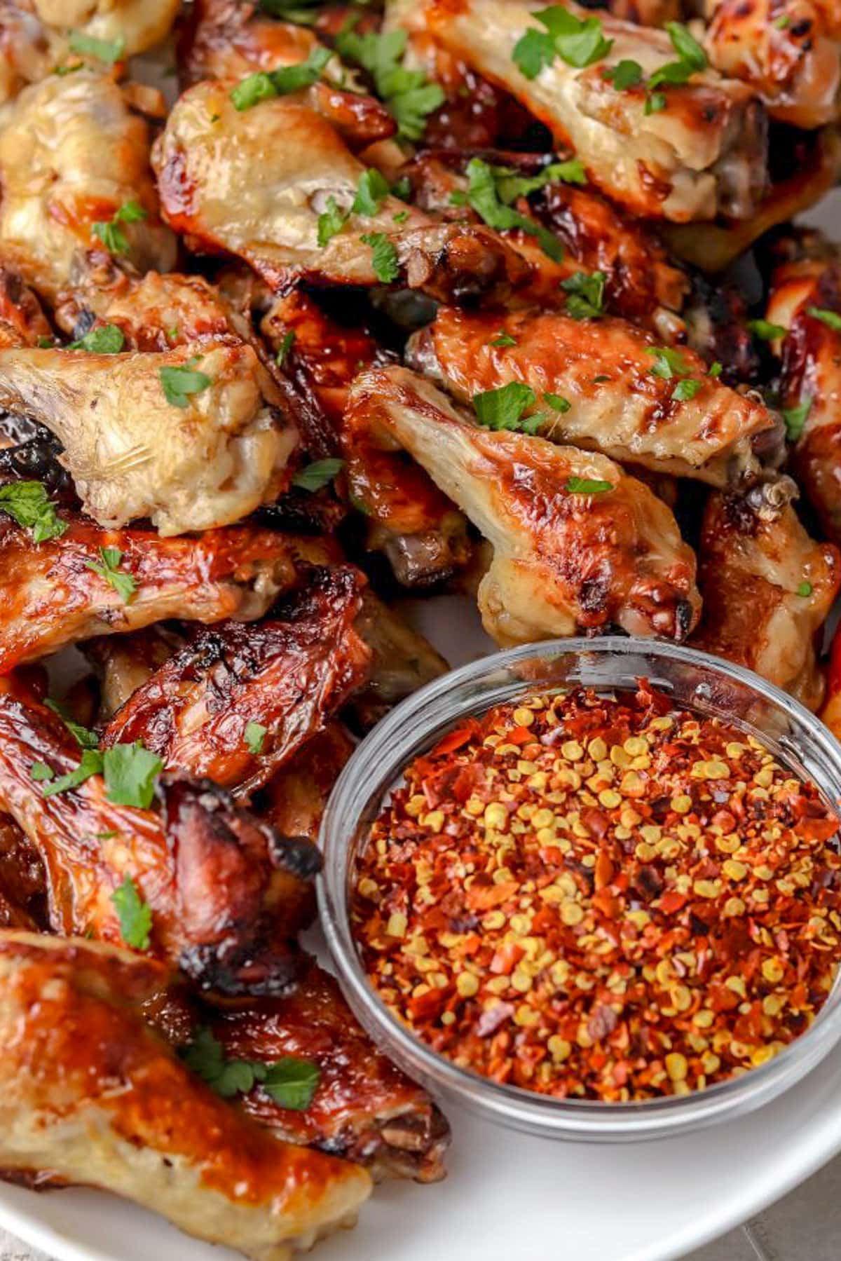 Juicy brined and cooked chicken wings piled on a platter next to a glass serving bowl of sauce with red pepper flakes.