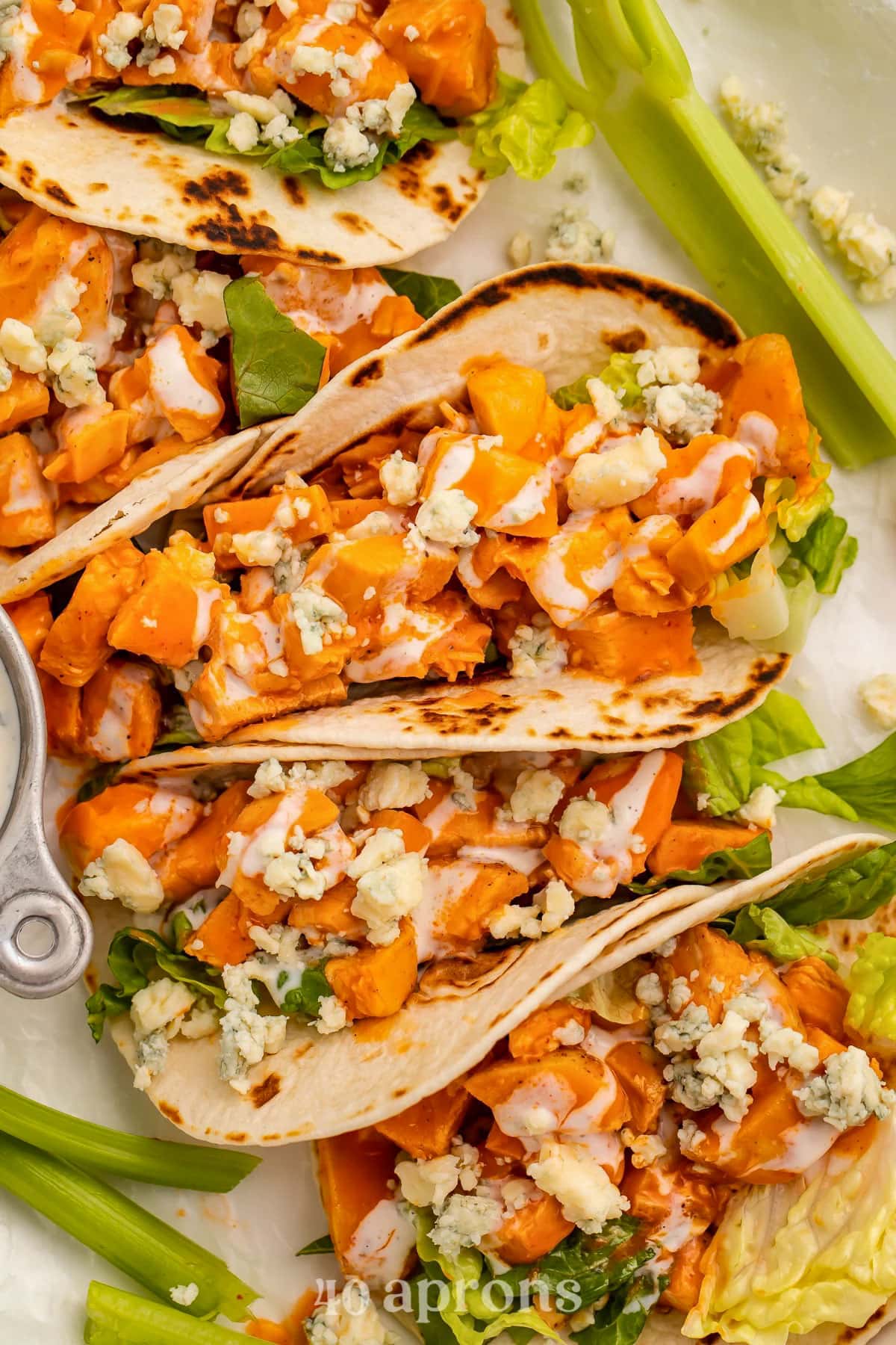 Top-down look at buffalo chicken in flour tortillas with crumbled cheese lined up on a platter with celery sticks.
