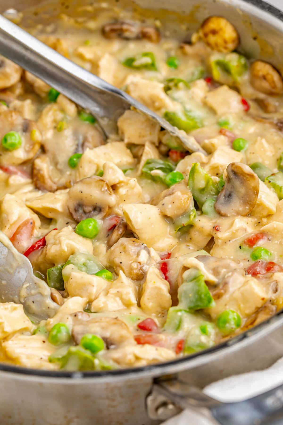Creamy chicken a la king in a large silver skillet with bright green peas and a pair of silver tongs.