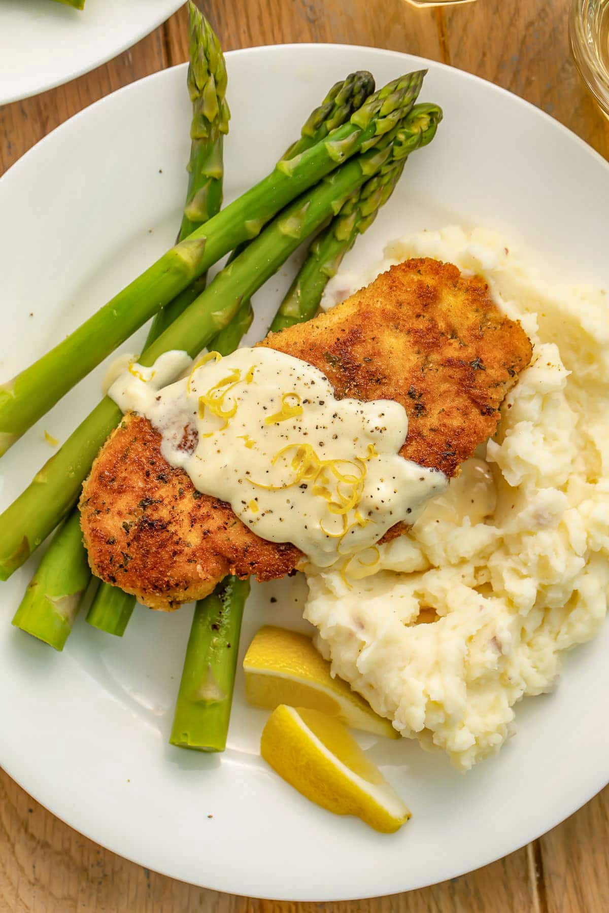 Copycat chicken costoletta on a white plate with mashed potatoes, green asparagus, and a creamy sauce.