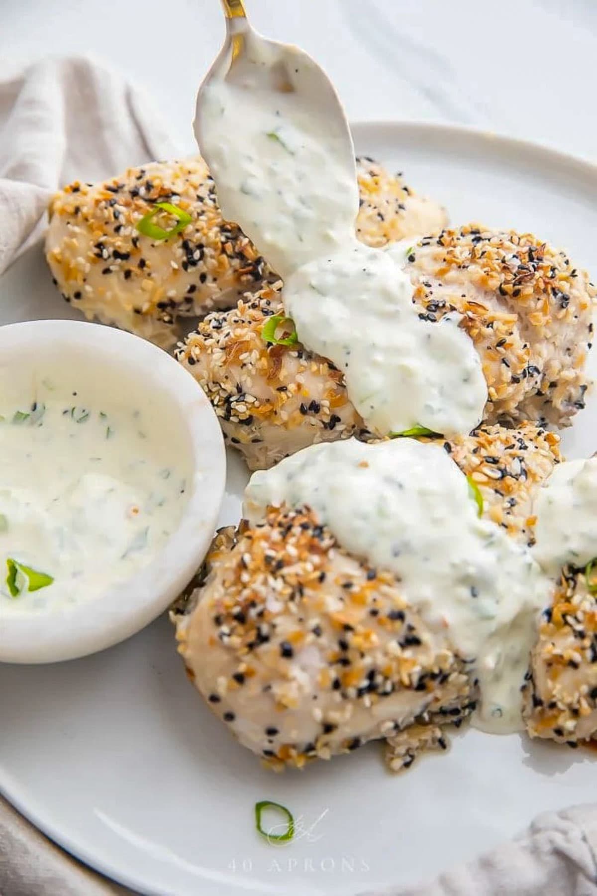 A spoonful of creamy scallion sauce being dolloped onto an everything bagel chicken breast on a plate.