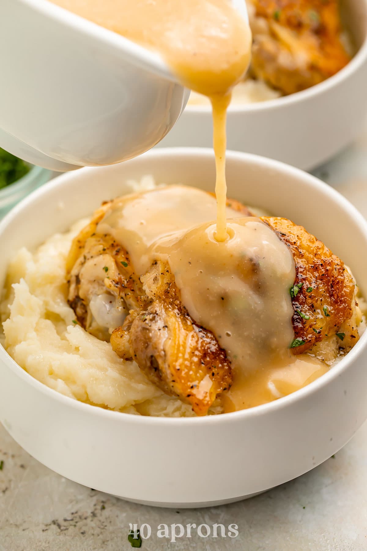 A chicken thigh, cooked in the Instant Pot, resting on top of mashed potatoes in a white bowl with gravy being poured over the chicken.