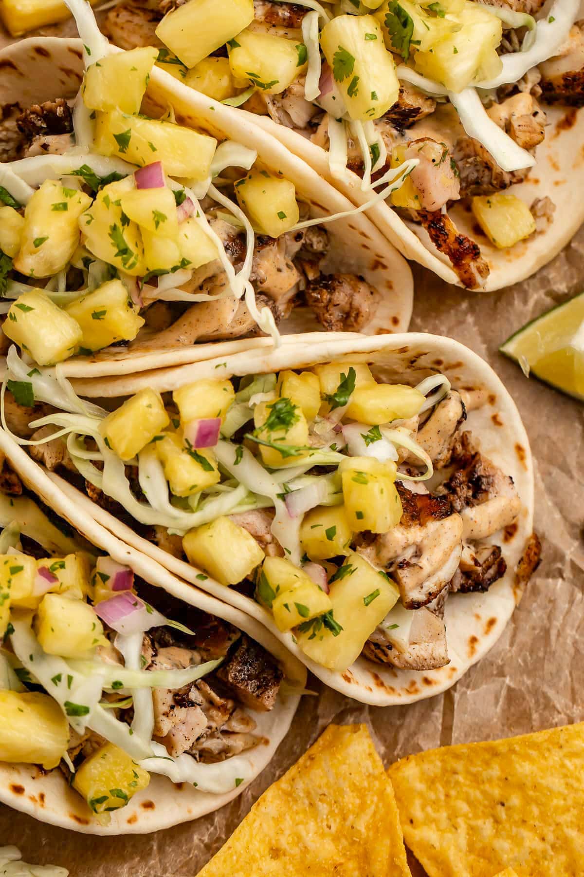 Overhead photo showing 4 jerk chicken tacos lined up on a sheet of parchment paper with tortilla chips and mango salsa.