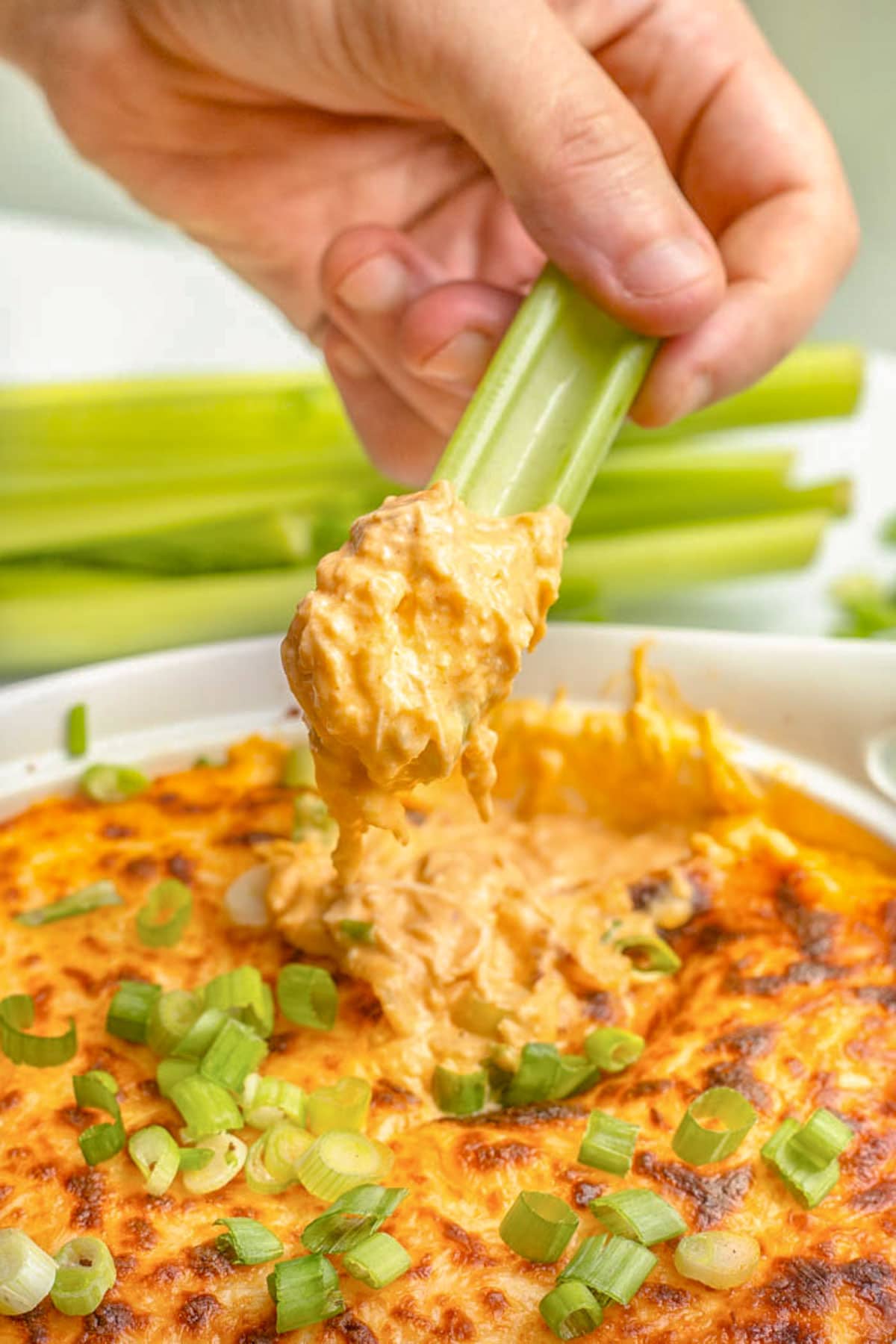 A white woman's hand holding a celery stick used to scoop keto buffalo chicken dip out of a bowl.