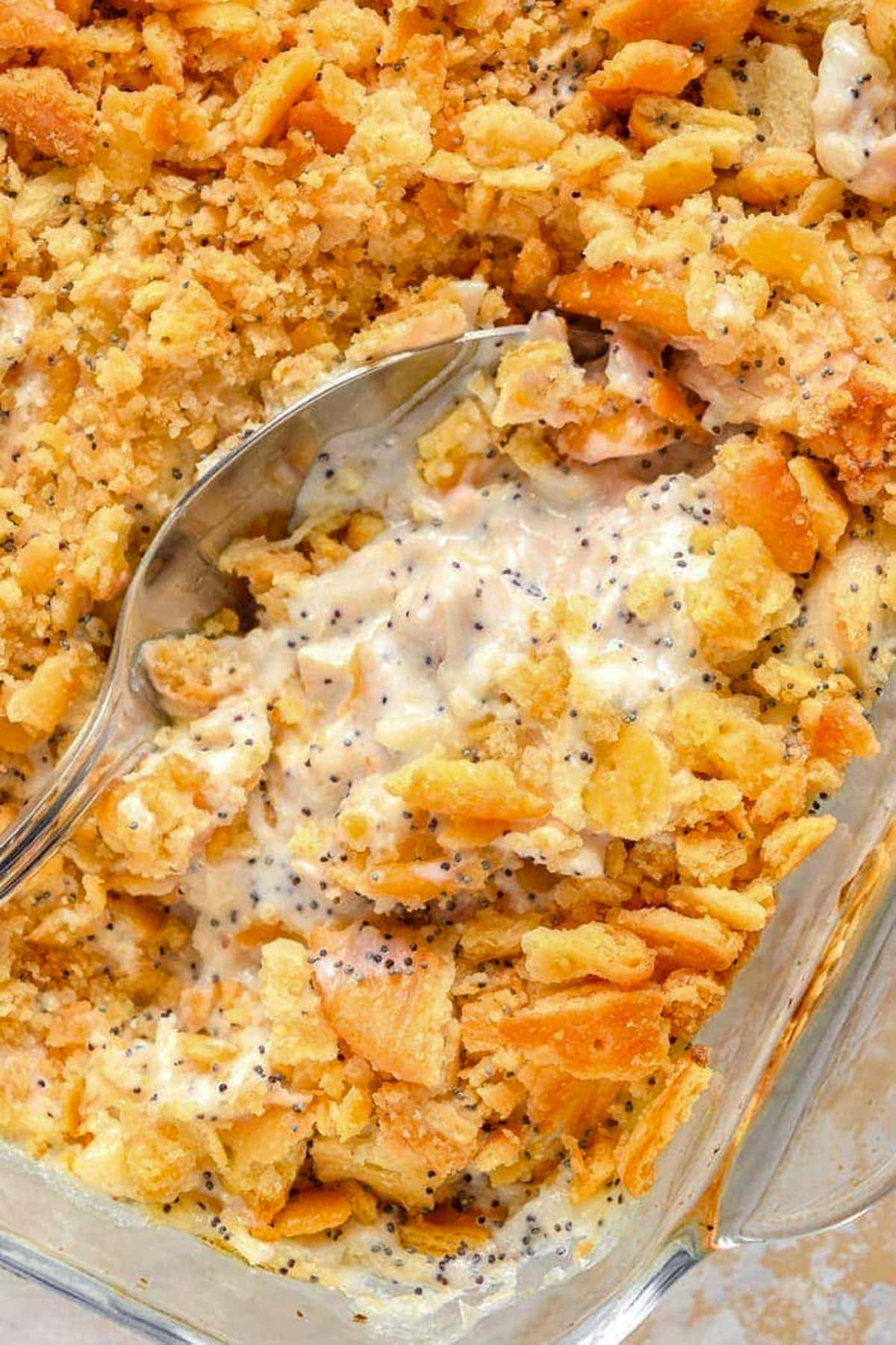 Poppy seed chicken casserole topped with Ritz crackers in a large dish, with a large spoon breaking through the crackers to show the creamy casserole beneath.