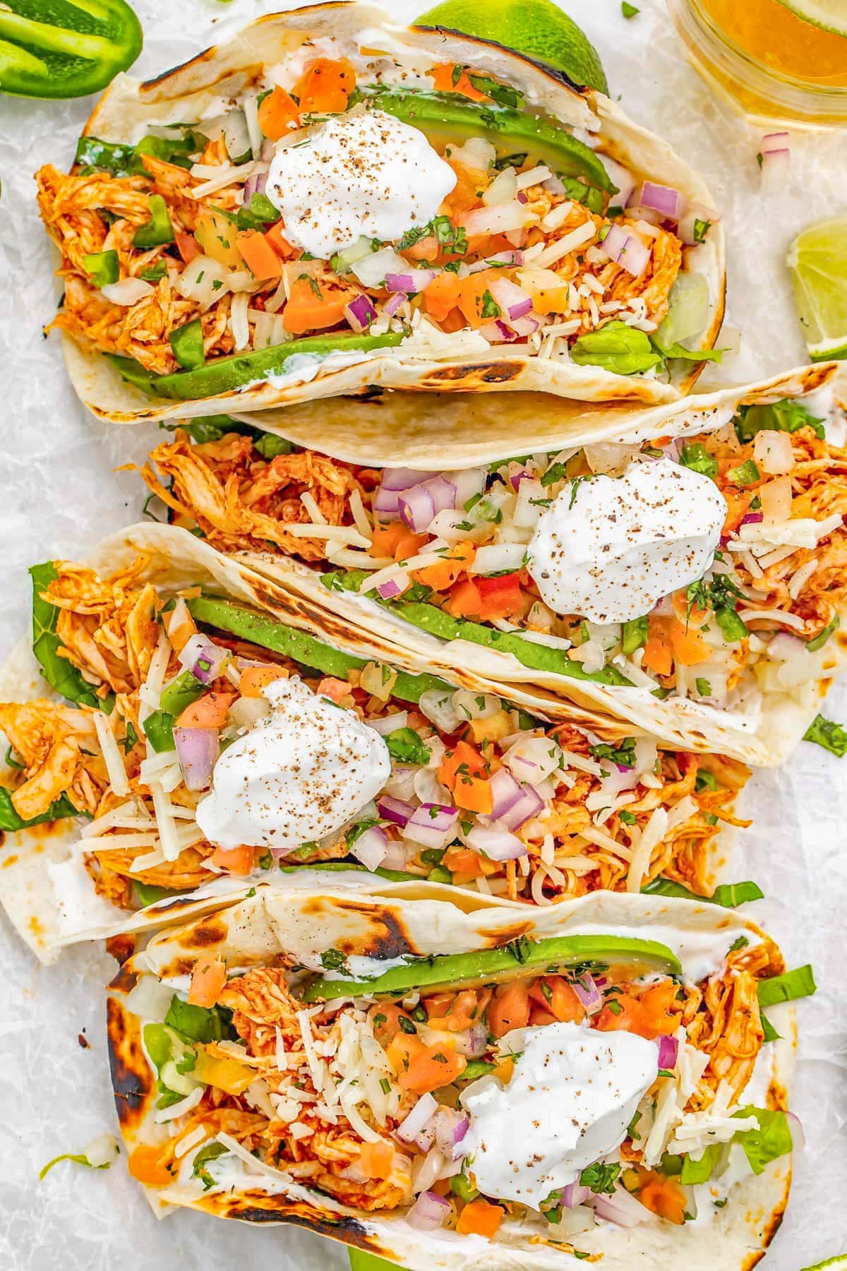 4 shredded chicken tacos topped with sour cream and toppings, placed side-by-side horizontally in a column on a sheet of parchment paper.